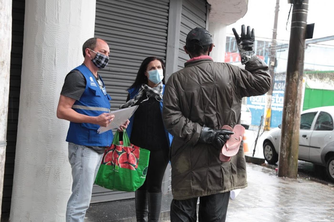 homem e mulher de equipe de rua e usando colete falam com morador em situação de rua. Ele está de costas. #paratodosverem