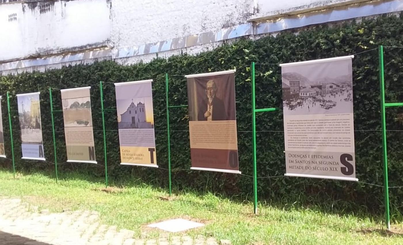 Paineis com imagens estão pendurados em sarrafos instalados em jardim. #paratodosverem 