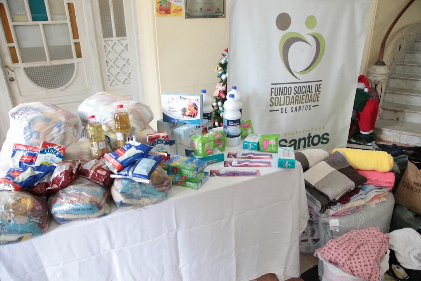 mesa com várias doações de alimentos e roupas empilhadas ao lado #paratodosverem