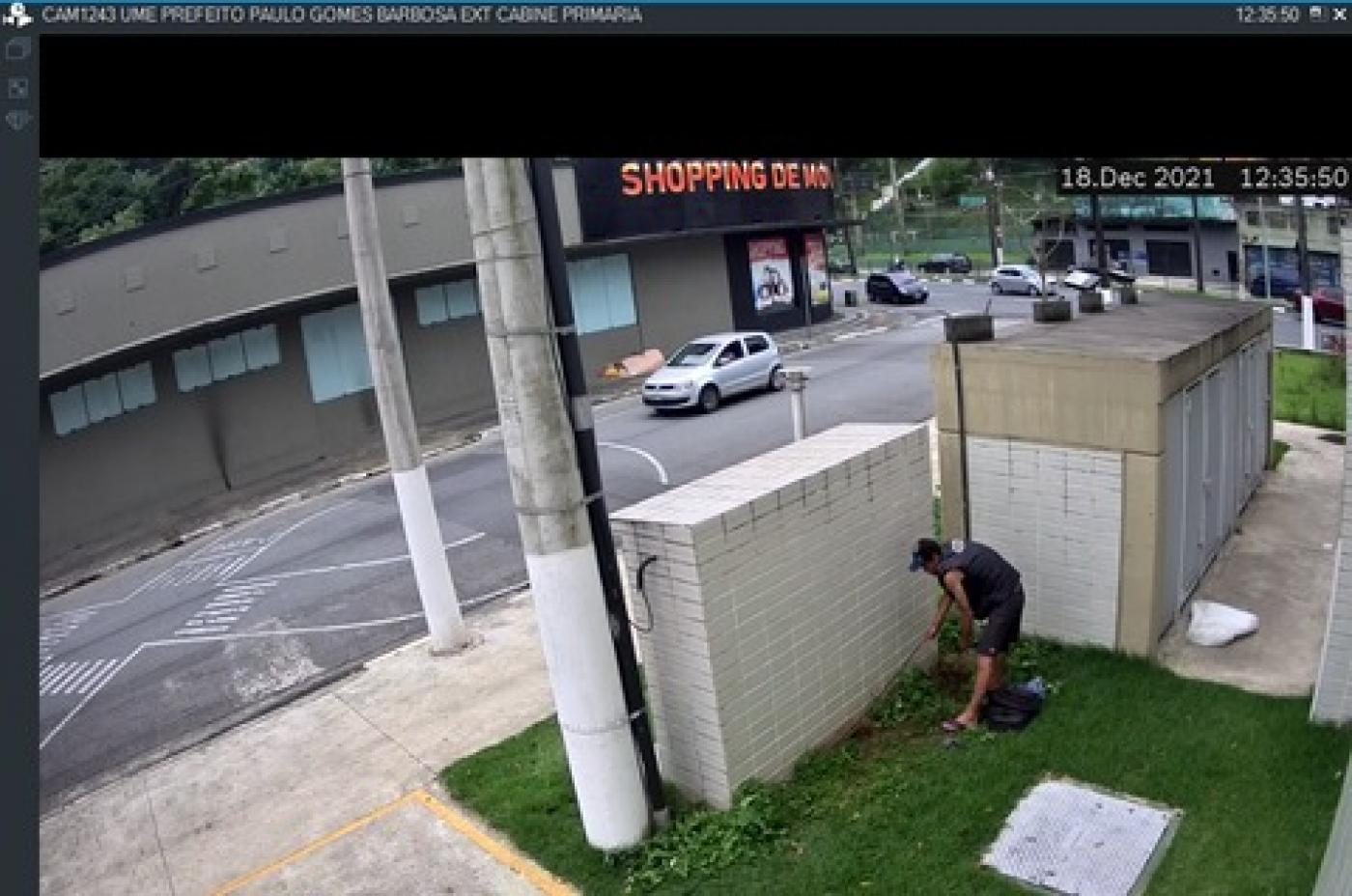 homem está puxando fio do solo junto a caixas de máquinas ao lado de posta.Imagem é de captação por câmera de monitoramento. #paratodosverem