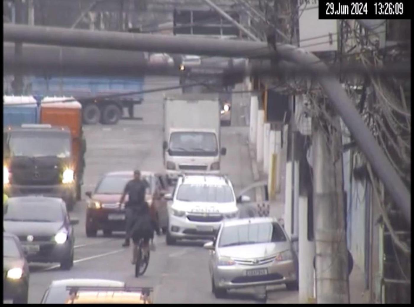 rua com carros circulando. Ao fundo, do lado direito,  uma viatura da PM, com um policial ao lado e uma bicicleta vindo à frente. #paratodosverem 