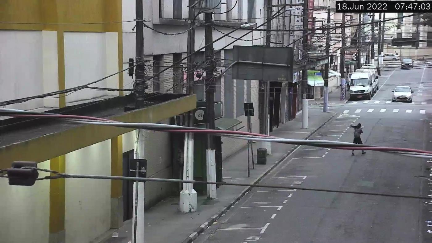 imagem a partir de câmera de monitoramento. No meio da rua se vê um homem caminhando e segurando peças nos ombros. #paratodosverem