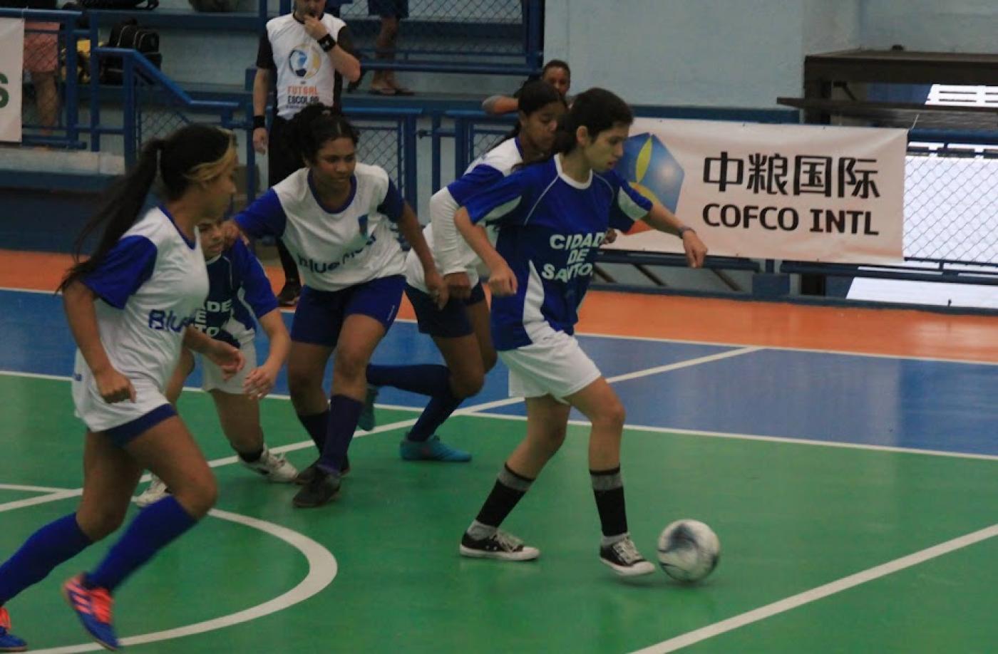 Meninas de uniformes branco e azul jogam futsal em quadra do Colégio Santista. #pratodosverem