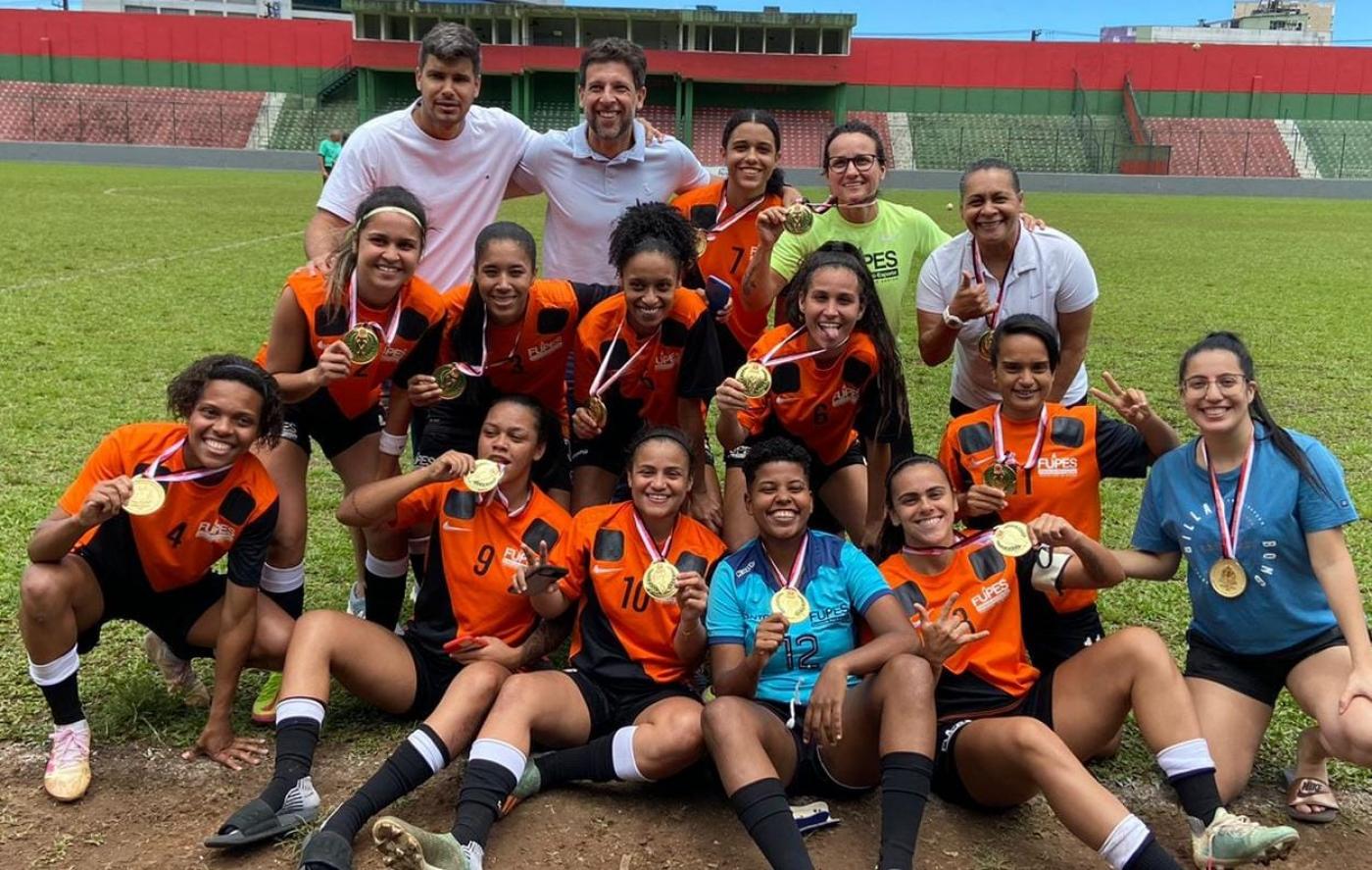 Jogadoras e comissão técnica mostram medalha com campo ao fundo #paratodosverem