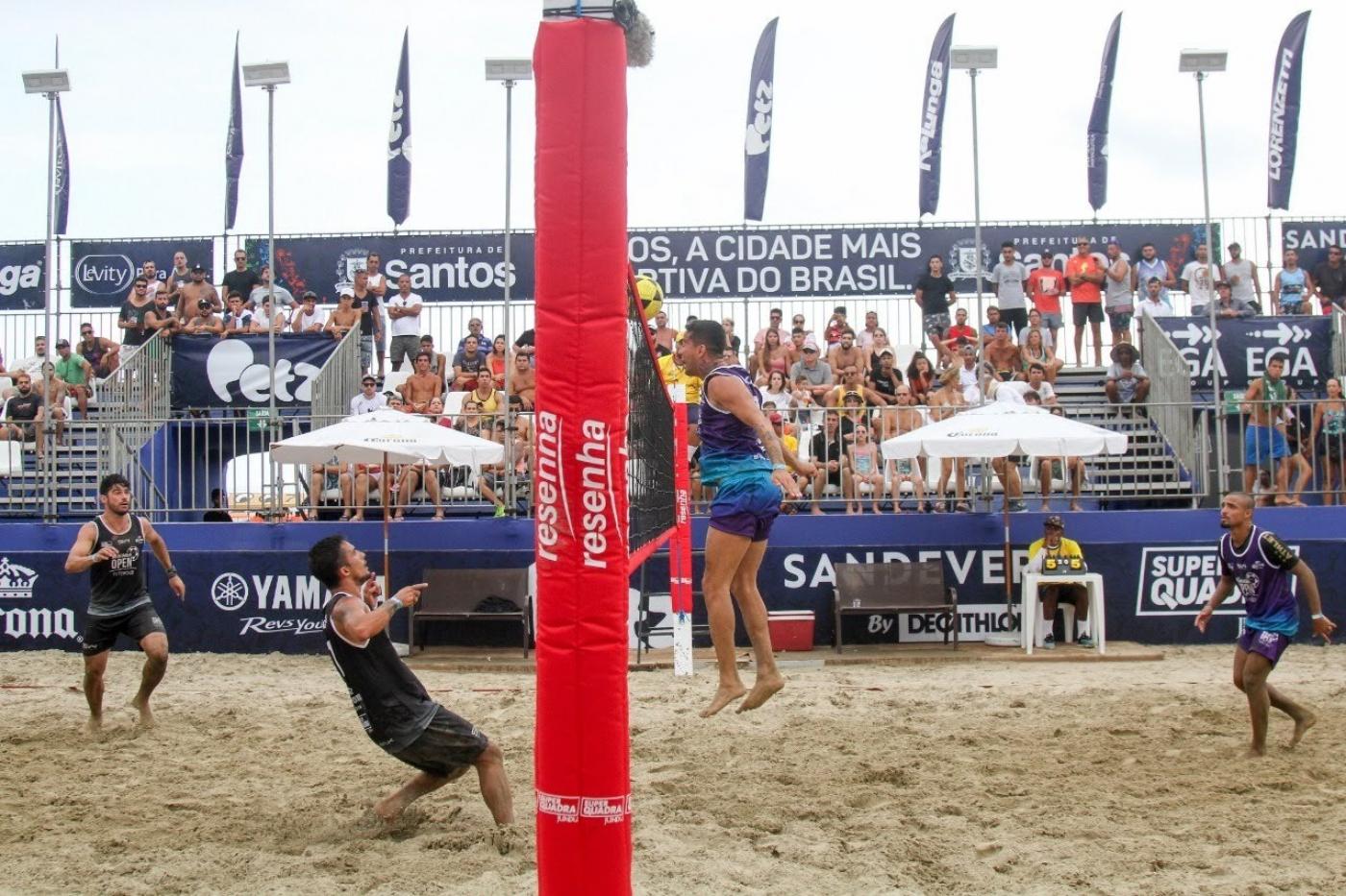 atletas estão jogando futvôlei na praia. Mastro da rede está ao centro da foto em primeiro plano. #paratodosverem