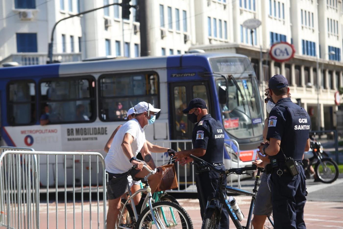 Dois guardas municipais abordam ciclista em ciclovia. Um ônibus passa atrás. #paratodosverem