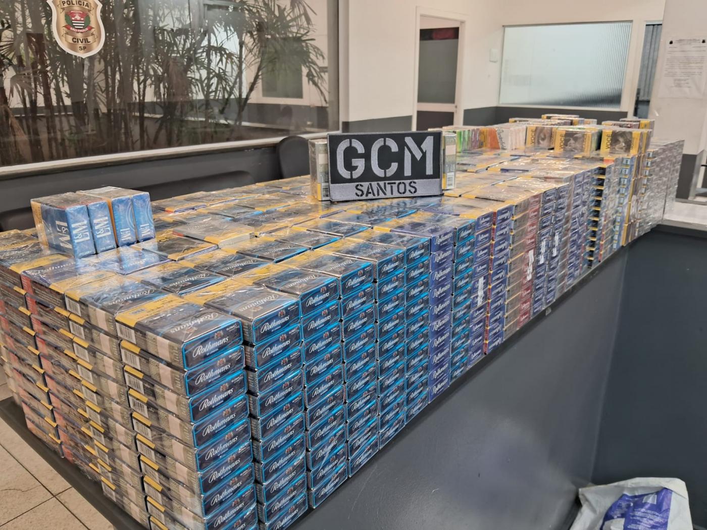 inúmeros pacotes com caixas de cigarro empilhados em sala. Placa com as letras gcm está sobre a pilha.  #paratodosverem