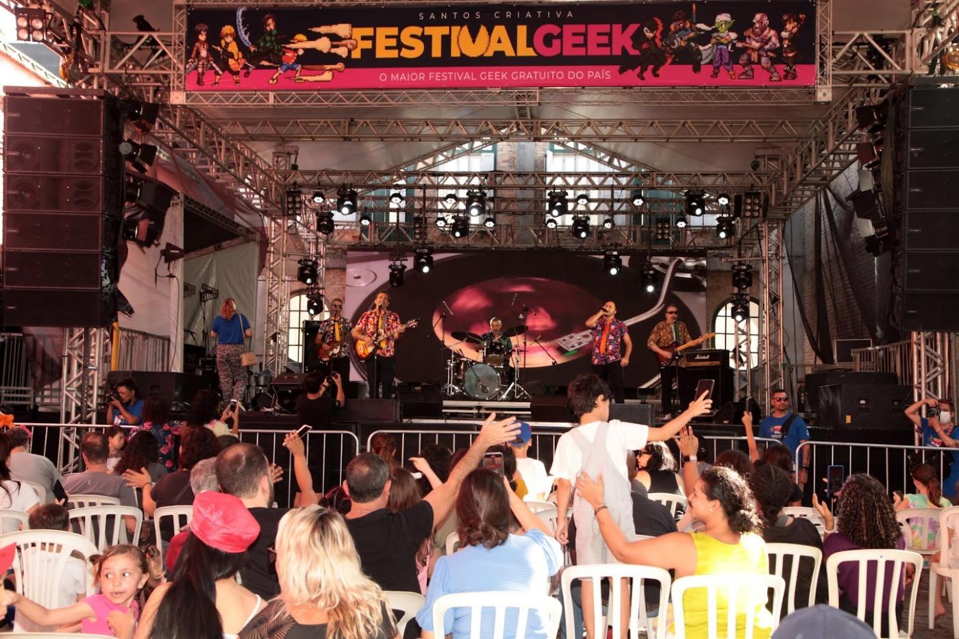 público sentado em cadeiras assistindo a show de músicos em palco. No alto do palco se lê Festival Geek. #paratodosverem