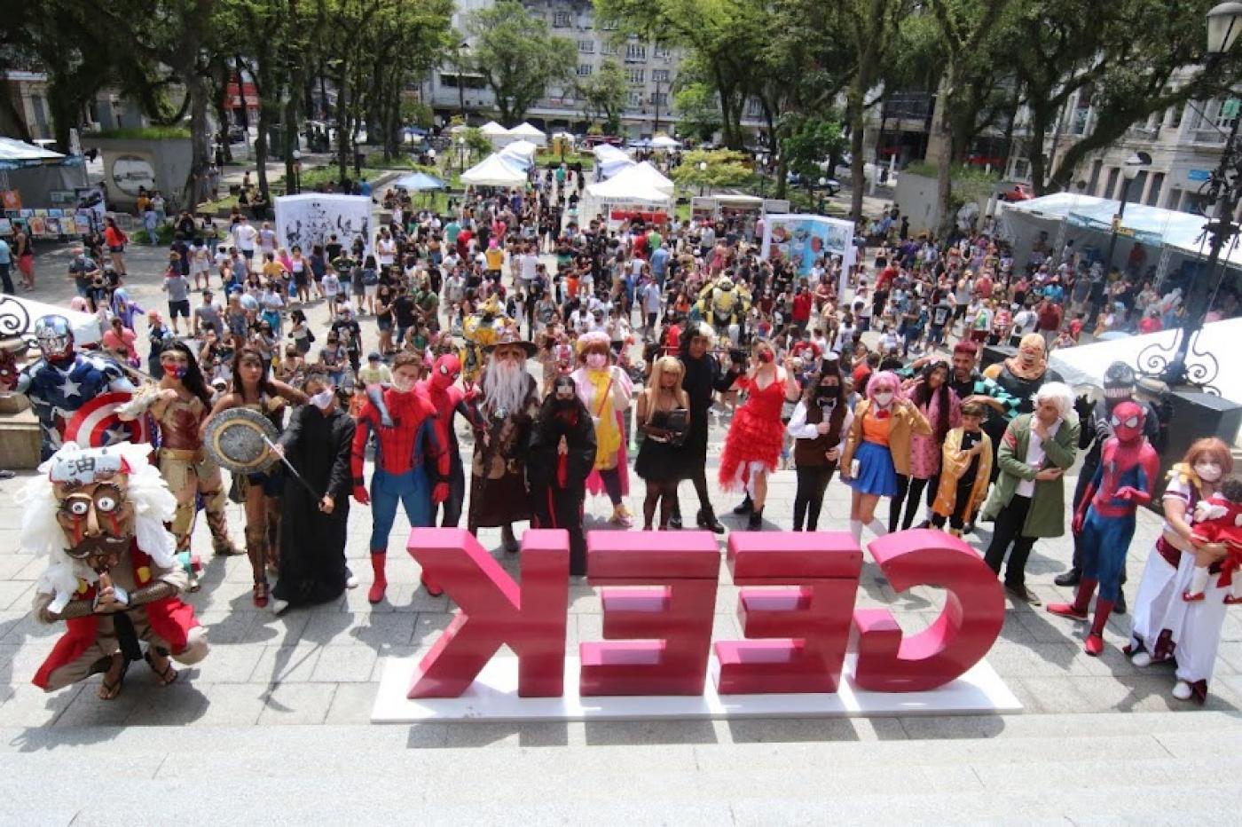 Cosplays ao lado de letreiro escrito geek e publico lotando a praça ao fundo #paratodosverem