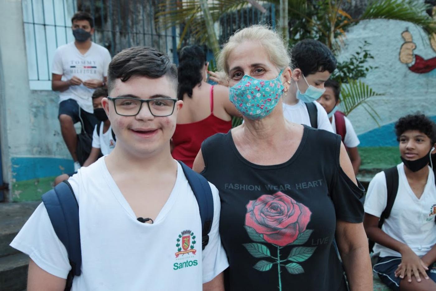 Giovanni ao lado da mãe na porta da escola. No fundo há outros estudantes uniformizados. #paratodosverem