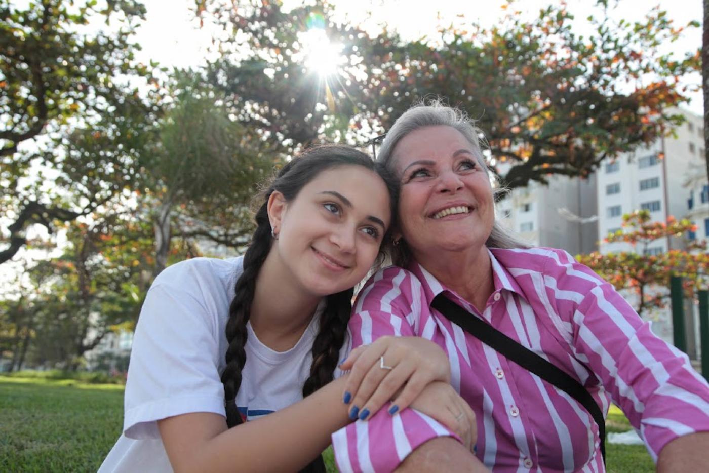 giulia entrelaça o braço direito da avó. Elas estão lado a lado, sentadas no jardim da praia. #paratodosverem. 