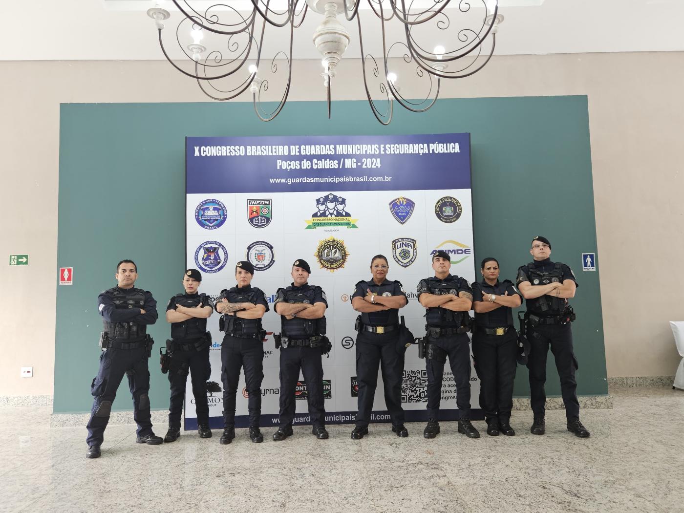 grupo de oito integrantes da guarda uniformizados e diante de um painel do evento. #paratodosverem