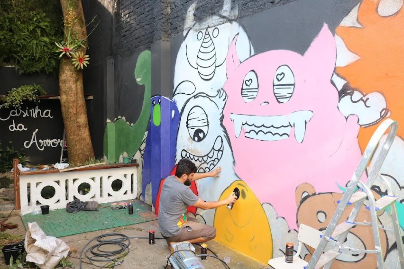 Artista, agachado, realiza pintura de monstrinho em parede de área interna da escola. #pracegover
