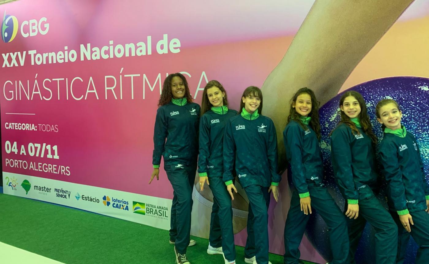 Seis meninas posam para foto na frente de outdoor do evento #paratodosverem