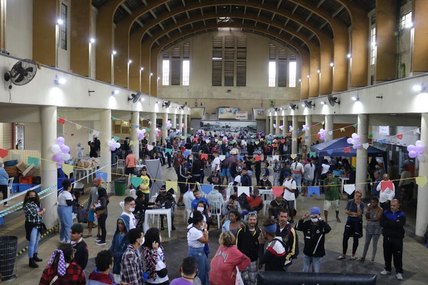 área interna do mercado muncipal lotada de pessoas. Bandeirinhas juninas decoram o ambiente penduradas entre as laterais do prédio. #paratodosverem