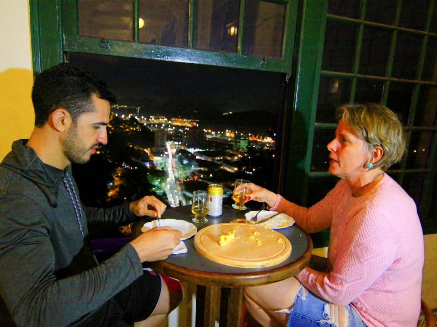 Homem e mulher comem e bebem ao lado de janela com vista de Santos a noite #paratodosverem