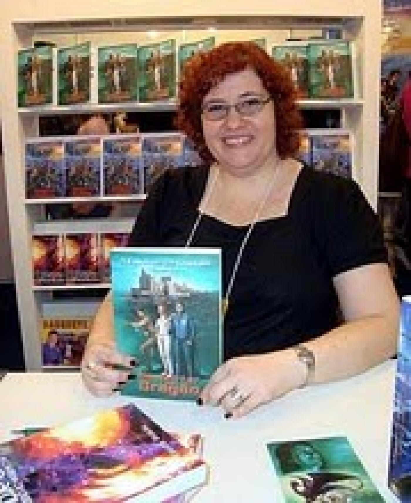 A escritora Helena Gomes está atrás de uma bancada segurando um livro. Ao fundo há uma estante com livros. #Pracegover
