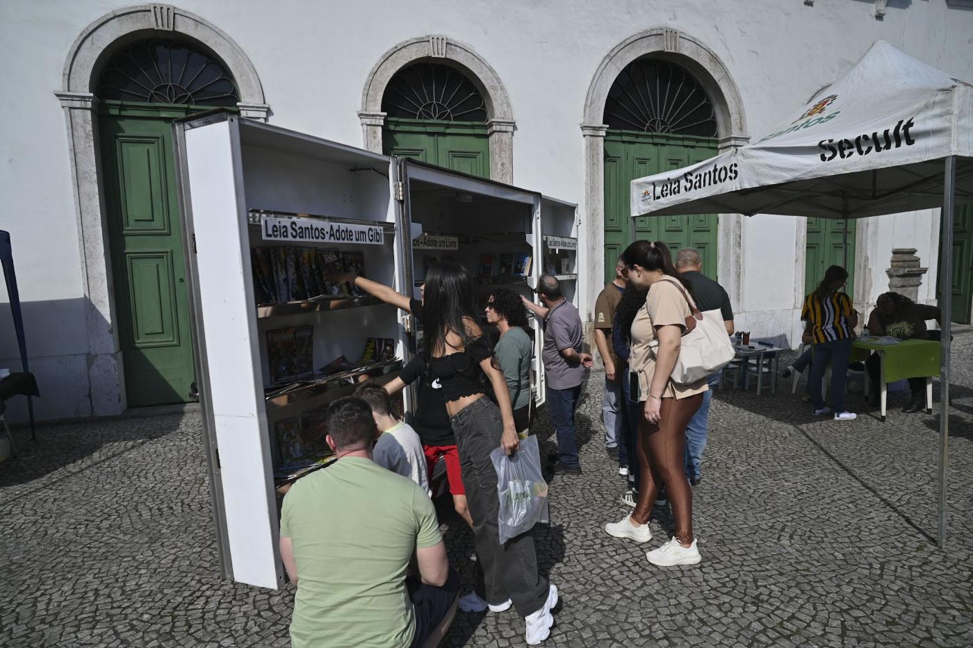 público em frente a estante de livros ao ar livre. #paratodosverem