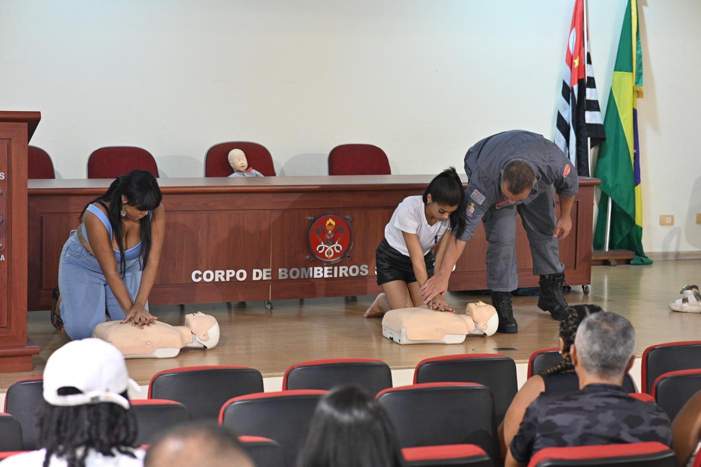 duas mulheres simulam massagem cardíaca em bonecos. militar dos bombeiros está ao lado de uma delas orientando. #paratodosverem