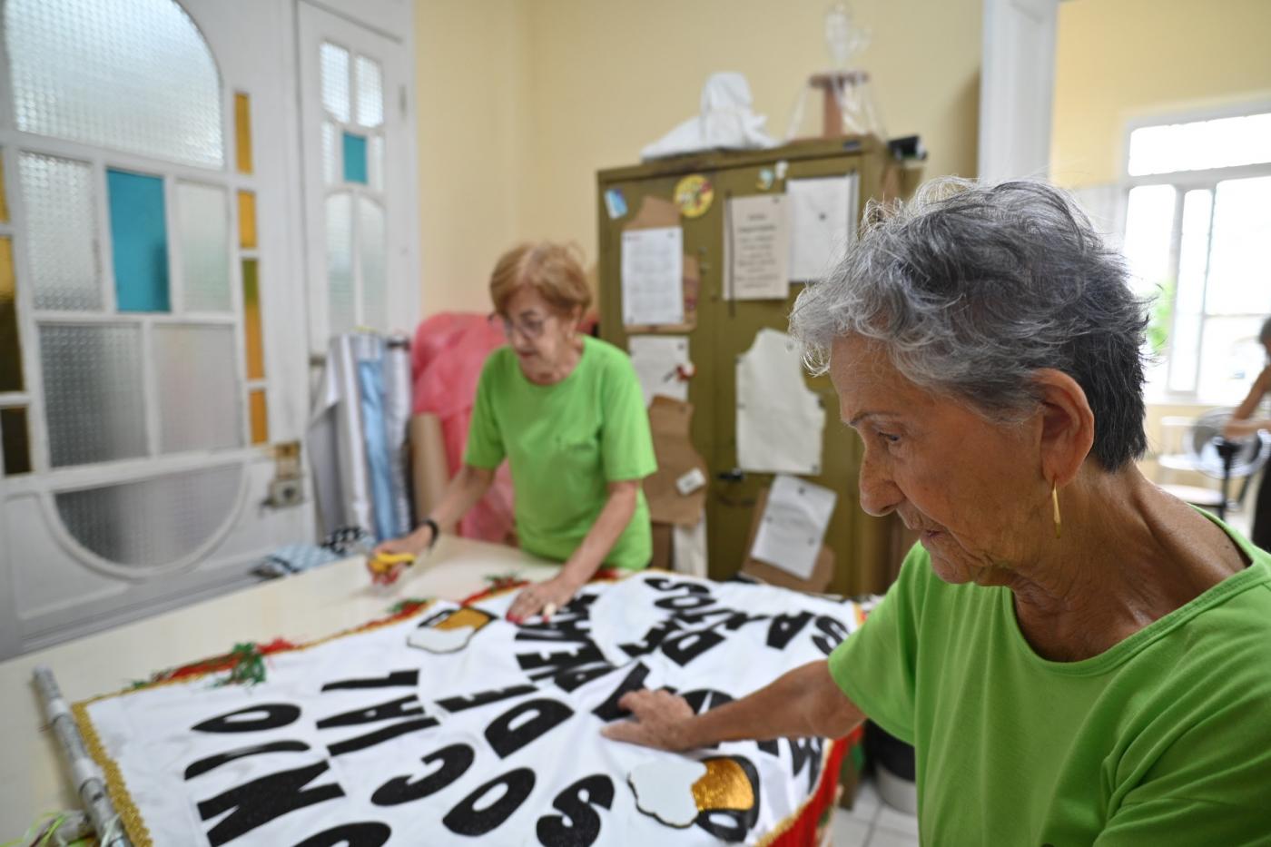 duas mulheres fazem costura em tecido com texto escrito. #paratodosverem