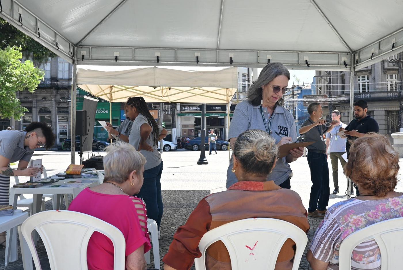 pessoas sentadas sob tenda armada em praça. #paratodosverem 