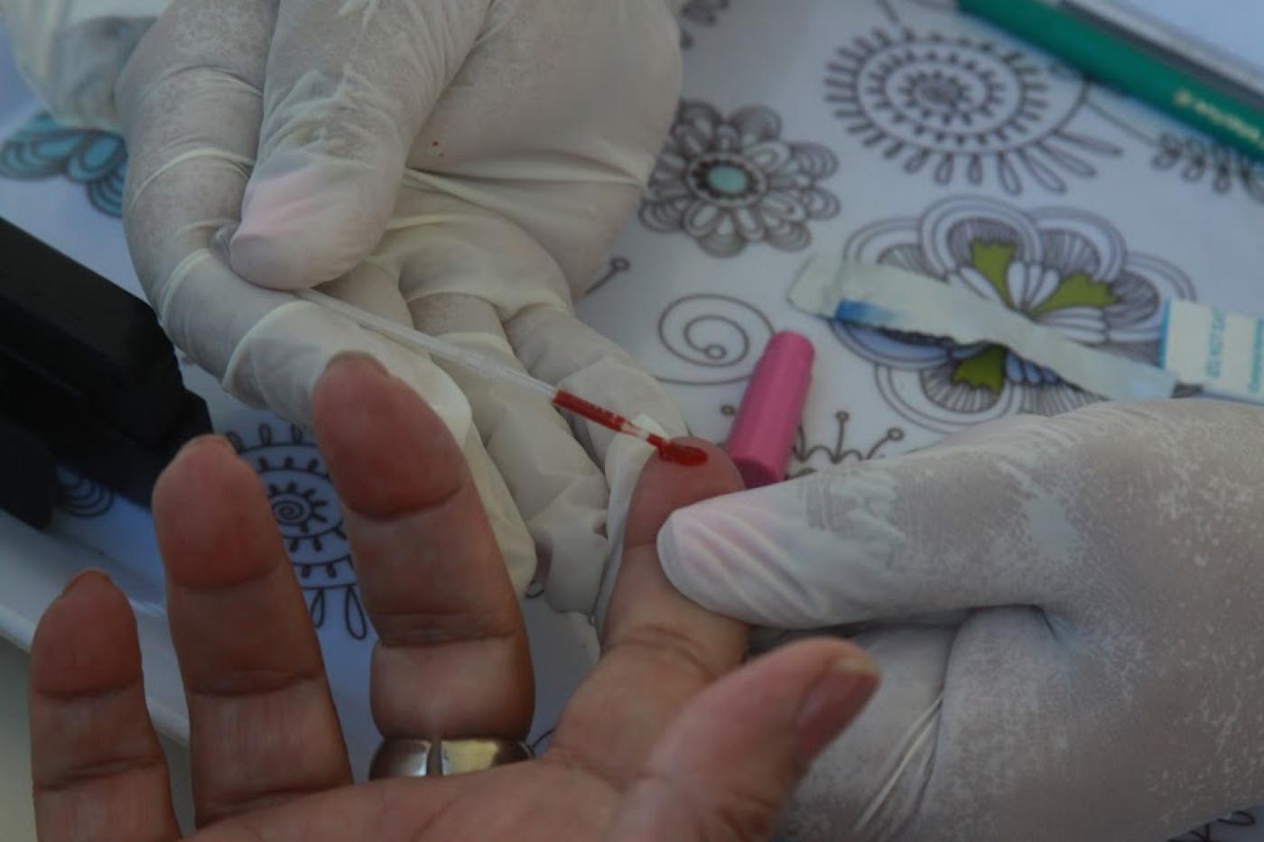 mãos com luvas usam objeto cortante para furar dedo de uma pessoa. Gota de sangue é colhida para exame. #paratodosverem 