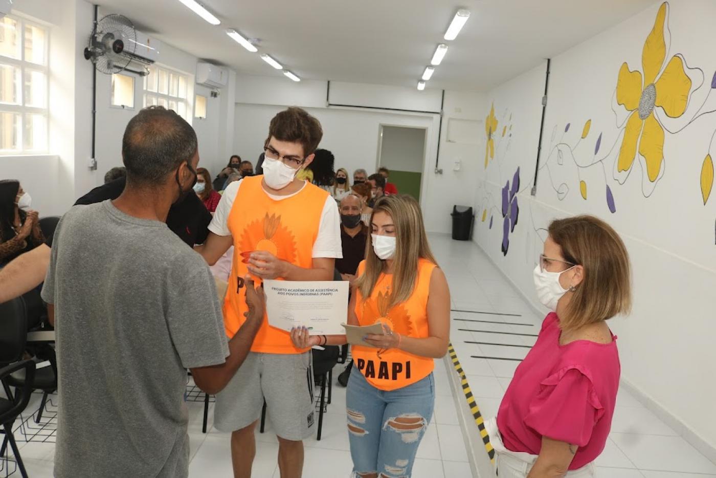 Dois voluntários usando camiseta de identificação recebem certificado de um homem que está de costas. A vice-prefeita está em pé, à direita. #paratodosverem