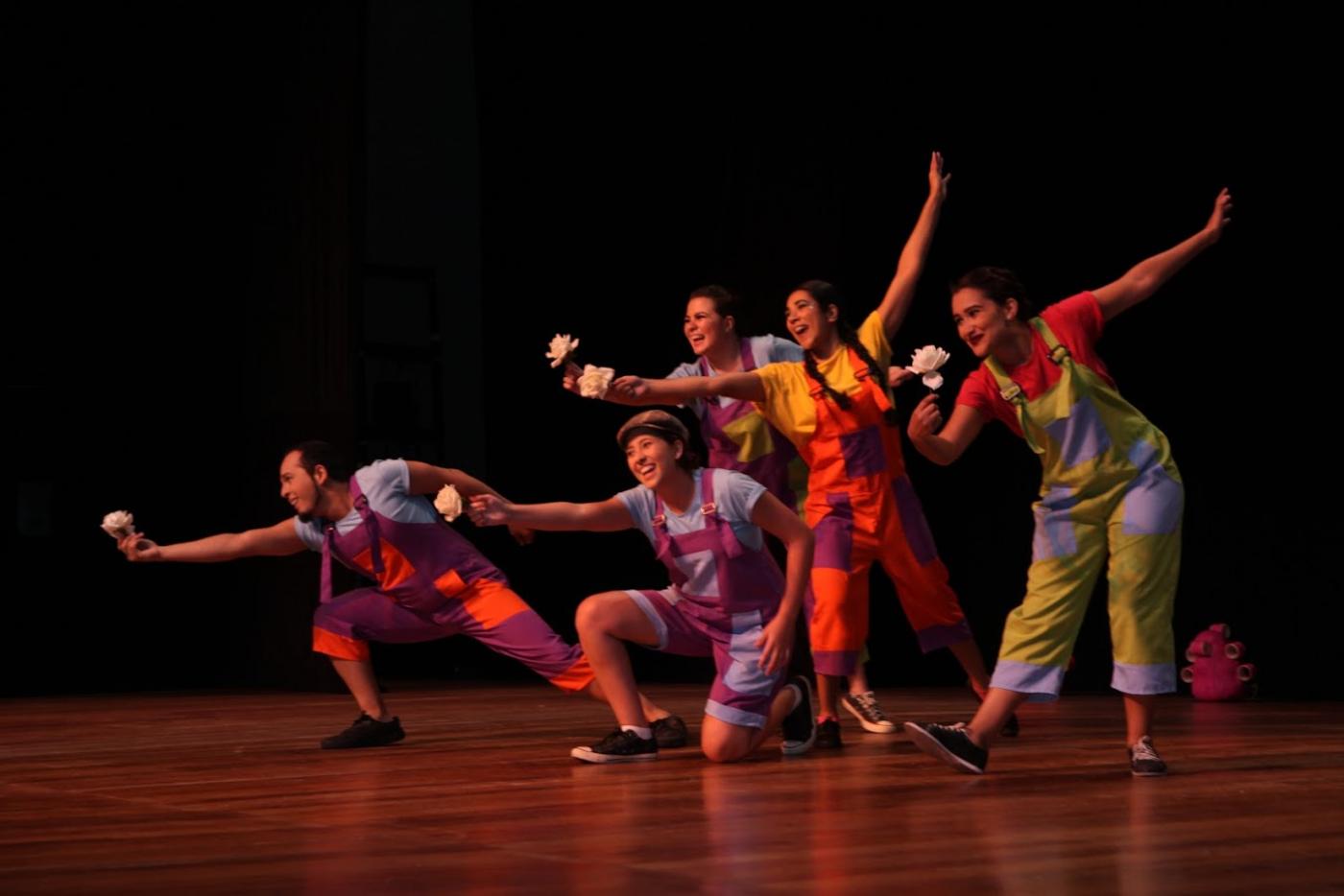 Artistas estão em palco de teatro, todos vestidos de palhaço e com flores nas mãos estendidas para a frente. #paratodosverem