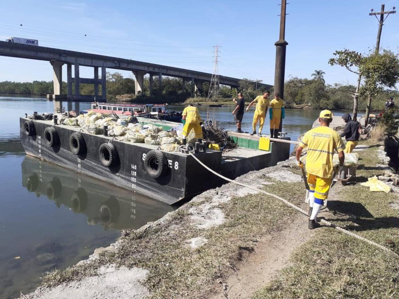 homens uniformizados colocam sacos de lixo em barcaça sobre a água. Ao fundo há uma ponte. Eles estão pisando num gramado sobre um pequeno cais à beira d'água. #paratodosverem