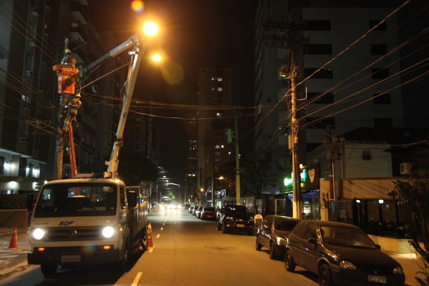 caminhão com braço basculante e cesto no topo está parado em rua para manutenção de iluminação no alto de um poste. #paratodosverem