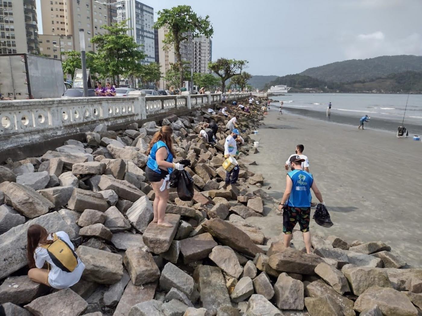 pessoas recolhem lixo em pedras na praia #paratodosverem