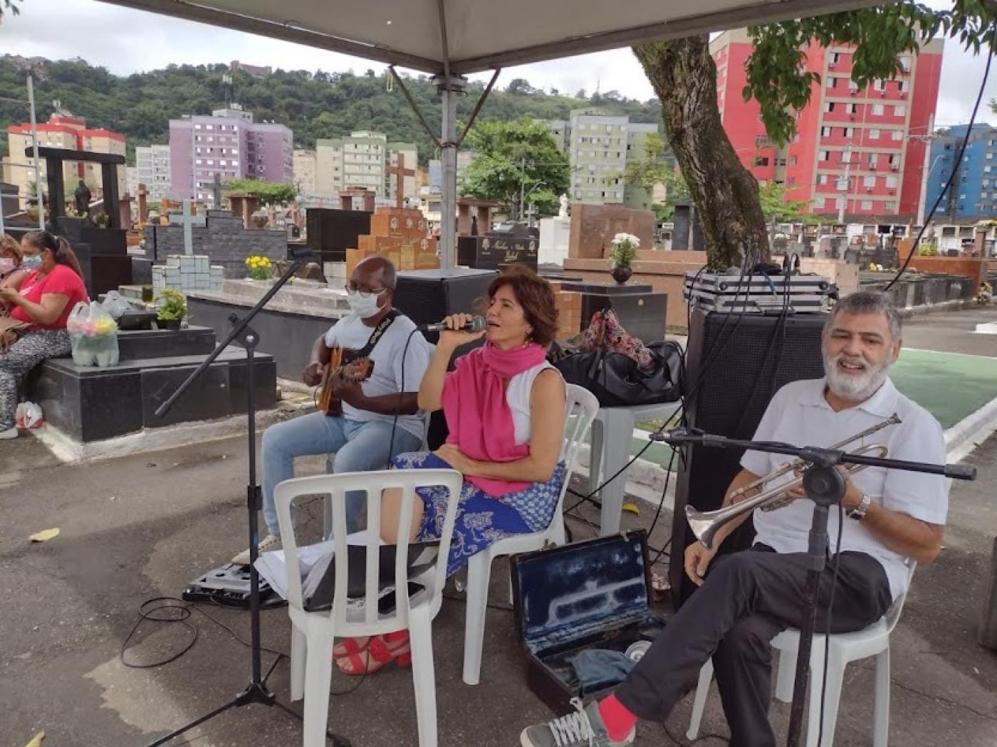 dois homens tocando instrumentos e uma mulher ao centro cantando, todos sob uma tenda, com campas ao fundo. #paratodosverem