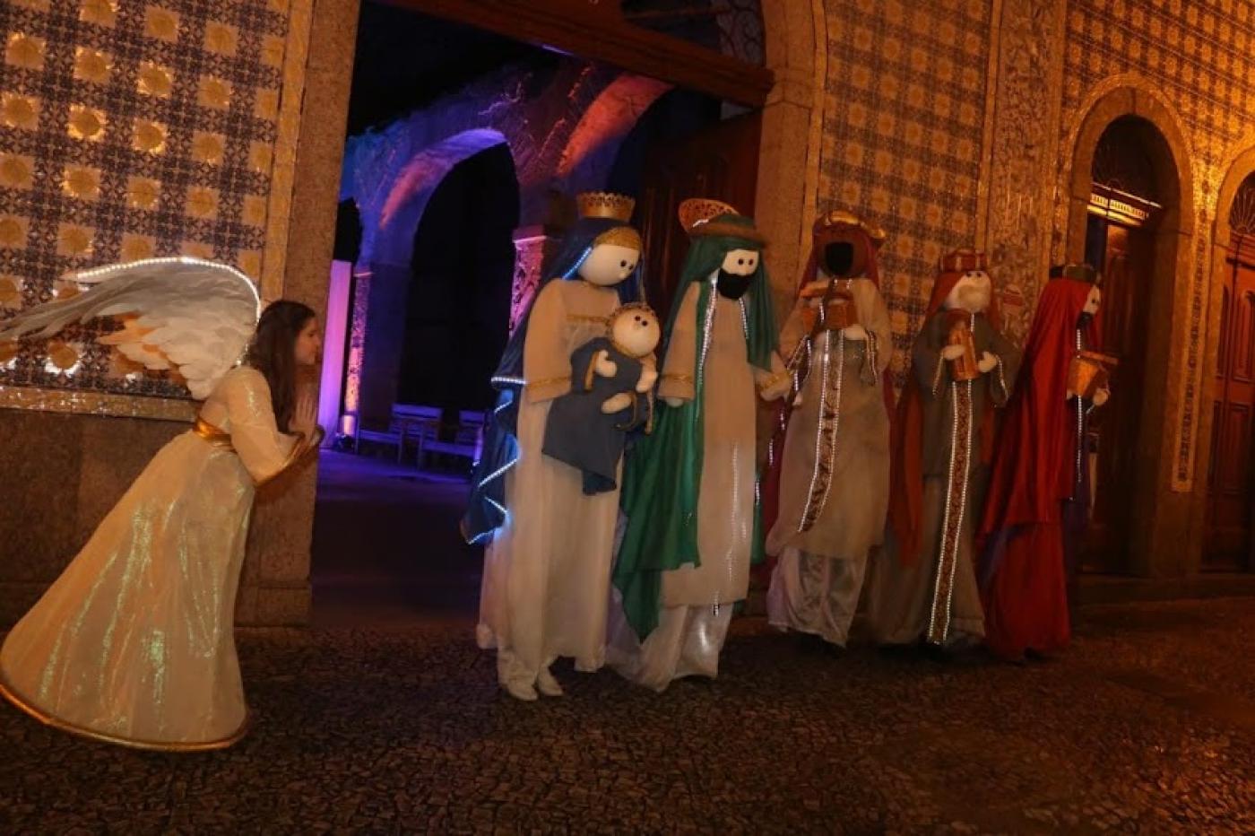 atores vestidos de um anjo, maria, josé e os três reis magos, além de um boneco de menino jesus estão á frente de uma casa de portas abertas. #paratodosverem 