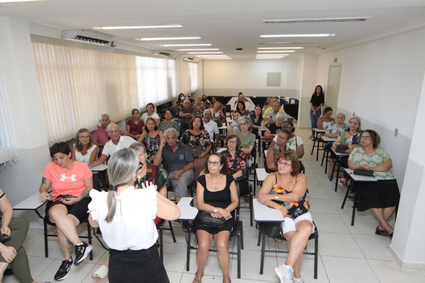 alunos estão sentados em sala de aula atentos a uma mulher que está falando. #paratodosverem