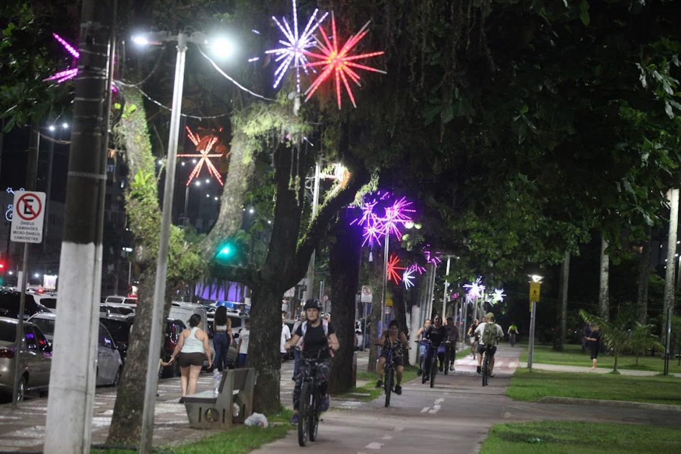 jardim da orla com árvores decoradas por luzes de natal. À direita passa a ciclovia, com bicicletas circulando. É noite. #paratodosverem 