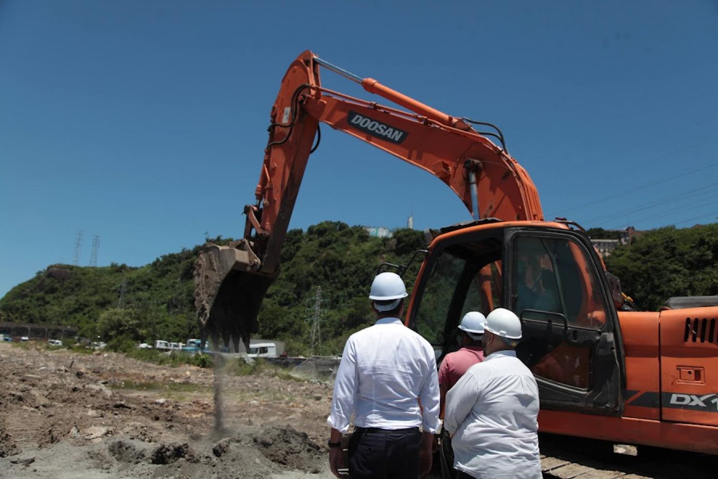 escavadeira opera em terreno. Três pessoas estão de costas para a foto usando capacete. #paratodosverem