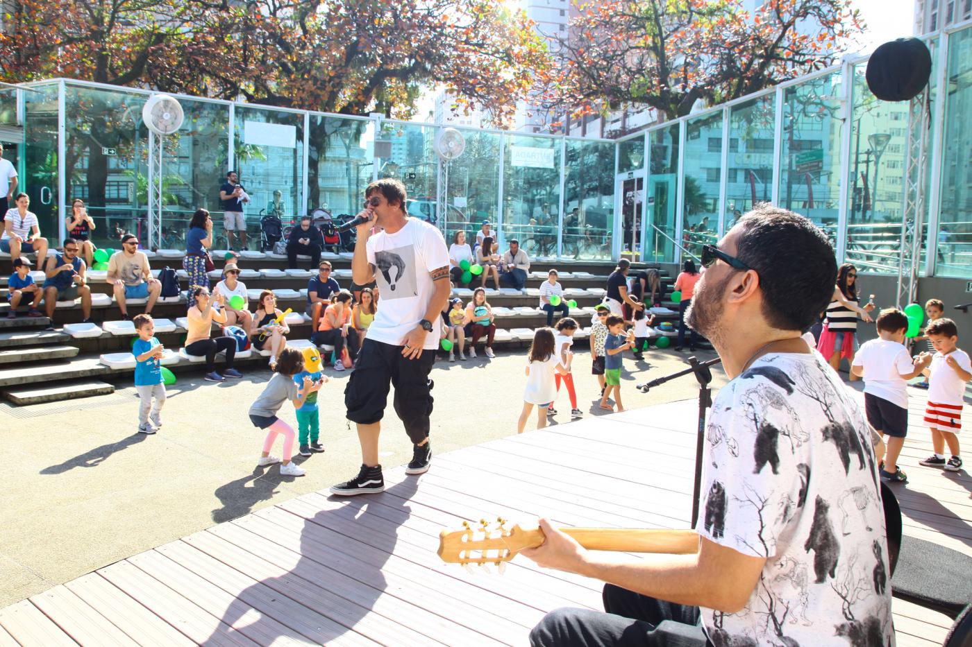 Com cerca de 20 crianças ao redor, homem canta enquanto outro toca guiterra no palco da Concha Acústica