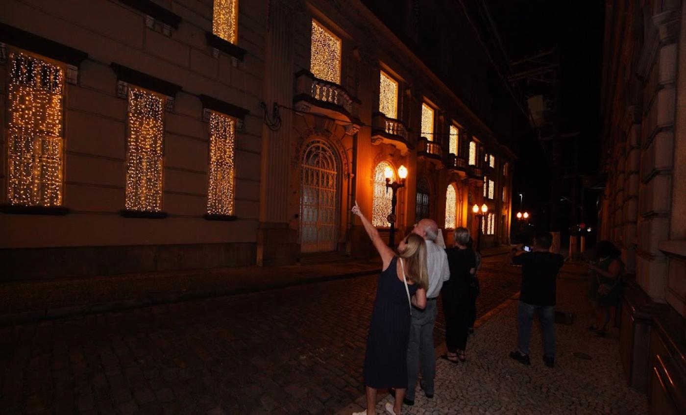 pessoas de um lado da calçada apontam para fachada de prédio iluminada para o natal. É noite. O prédio tem caráter histórico.  #paratodosverem 