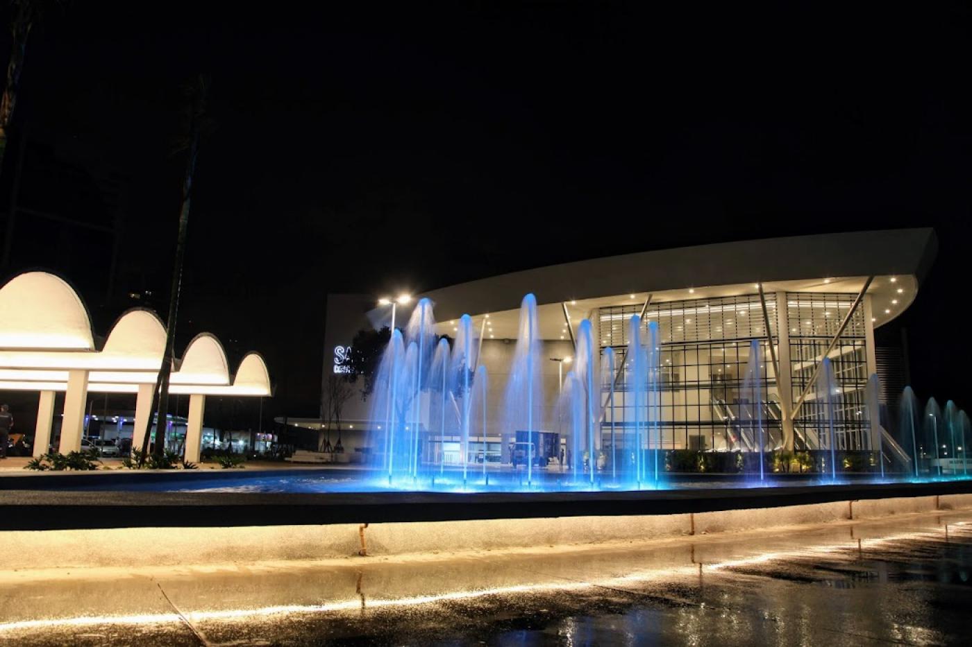 Foto noturna do centro de convenções com chafariz iluminado em azul a frente. #paratodosverem