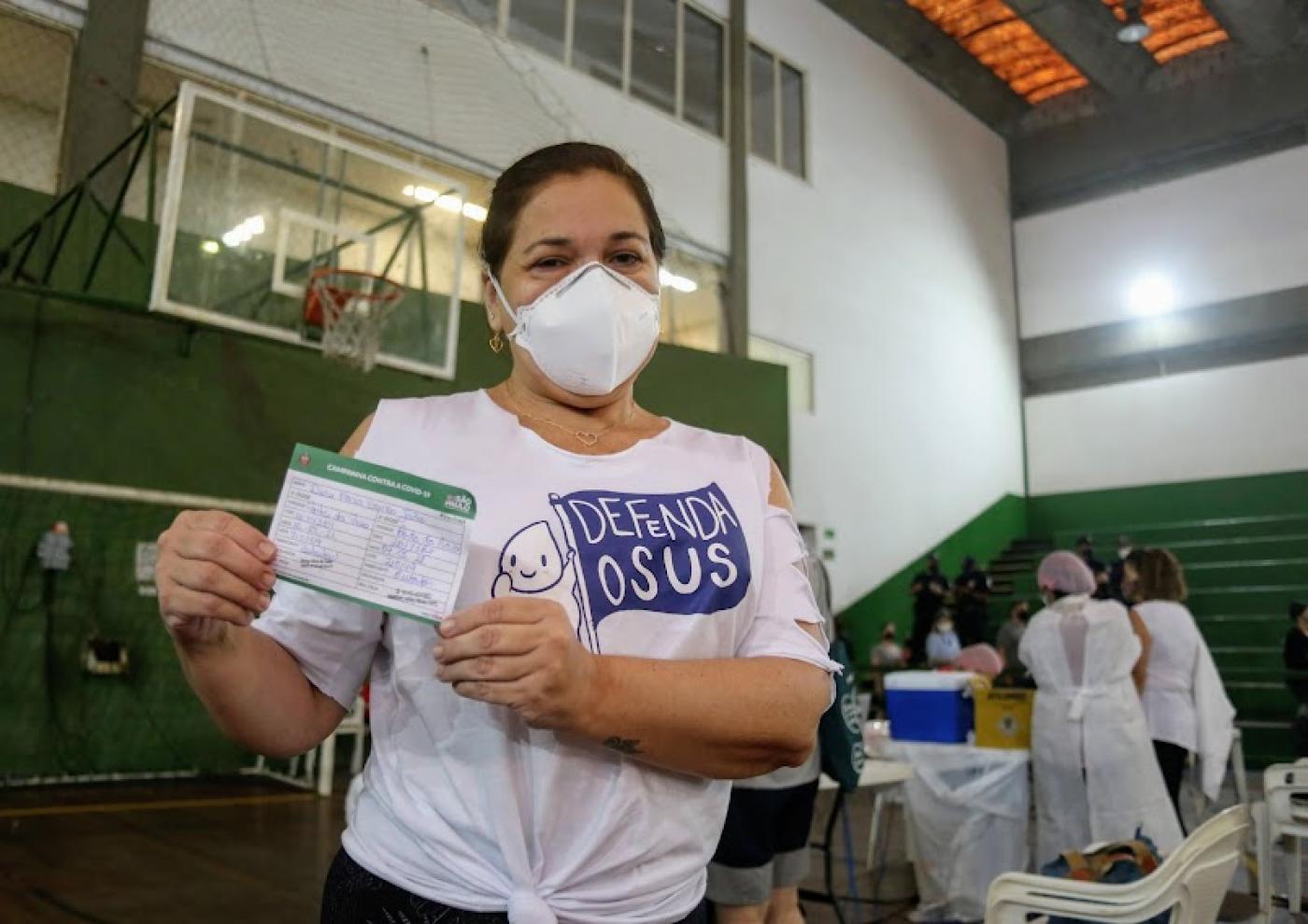 Mulher mostra carteira de vacinação, com enfermeiros ao fundo. #paratodosverem