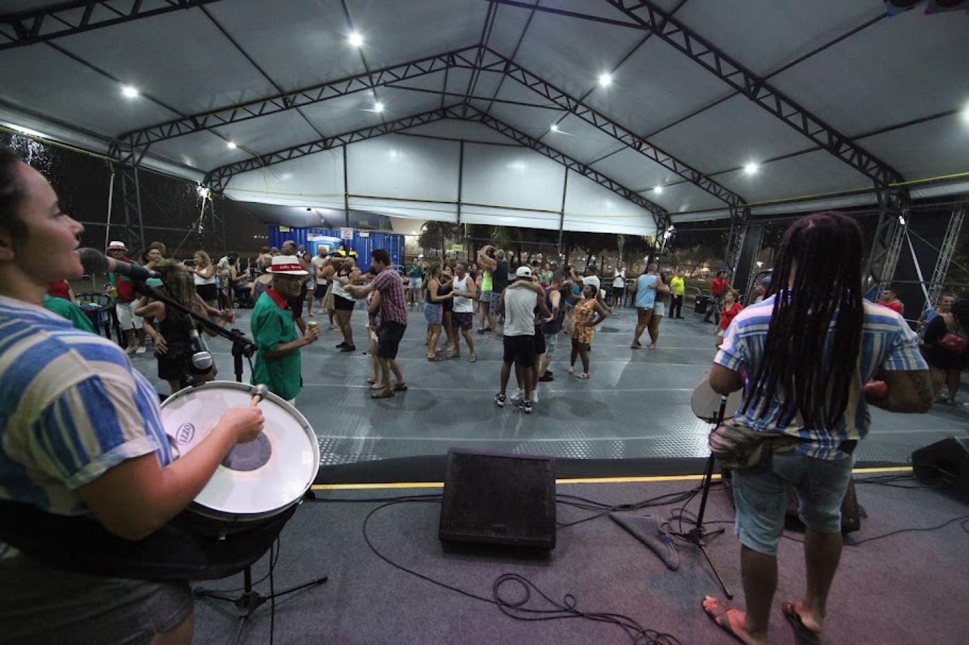 músicos tocam no palco enquanto público dança ao fundo sob tenda. #paratodosverem 