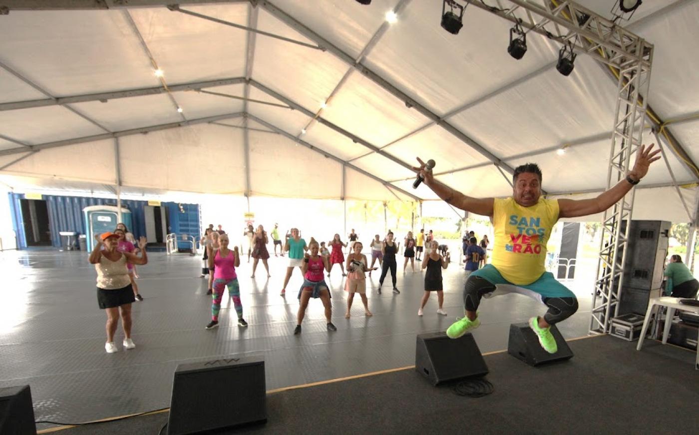 professor de dança dá salto no ar sobre o palco. Ao fundo, mulheres se movimentam. #paratodosverem 