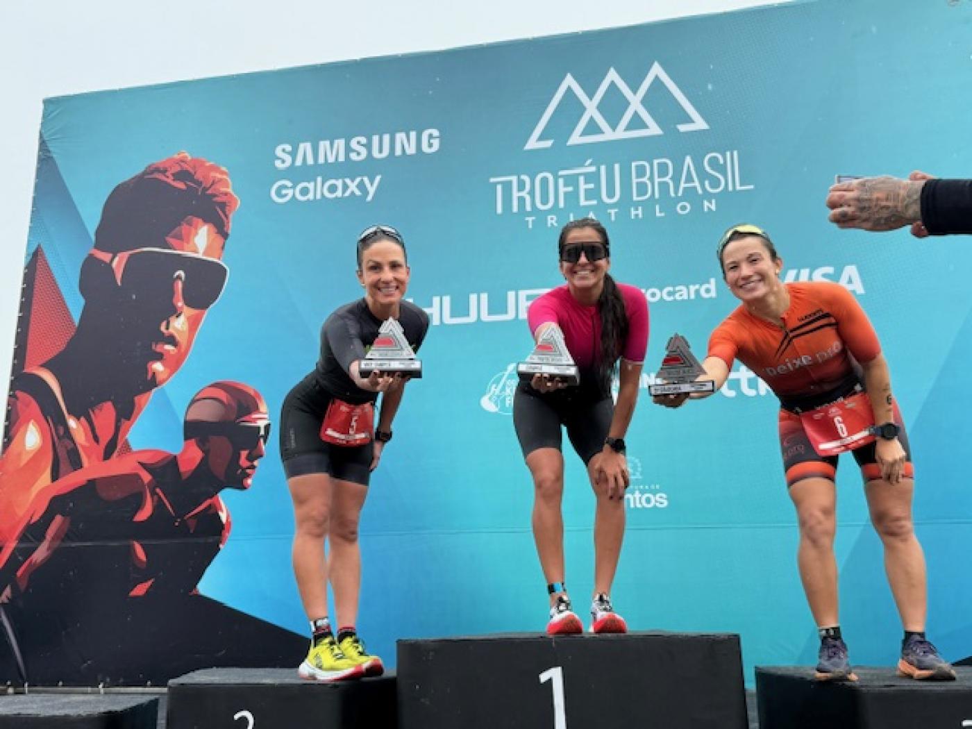 três mulheres no pódio seguram seus troféus e os apontam para a foto sorrindo. #paratodosverem 