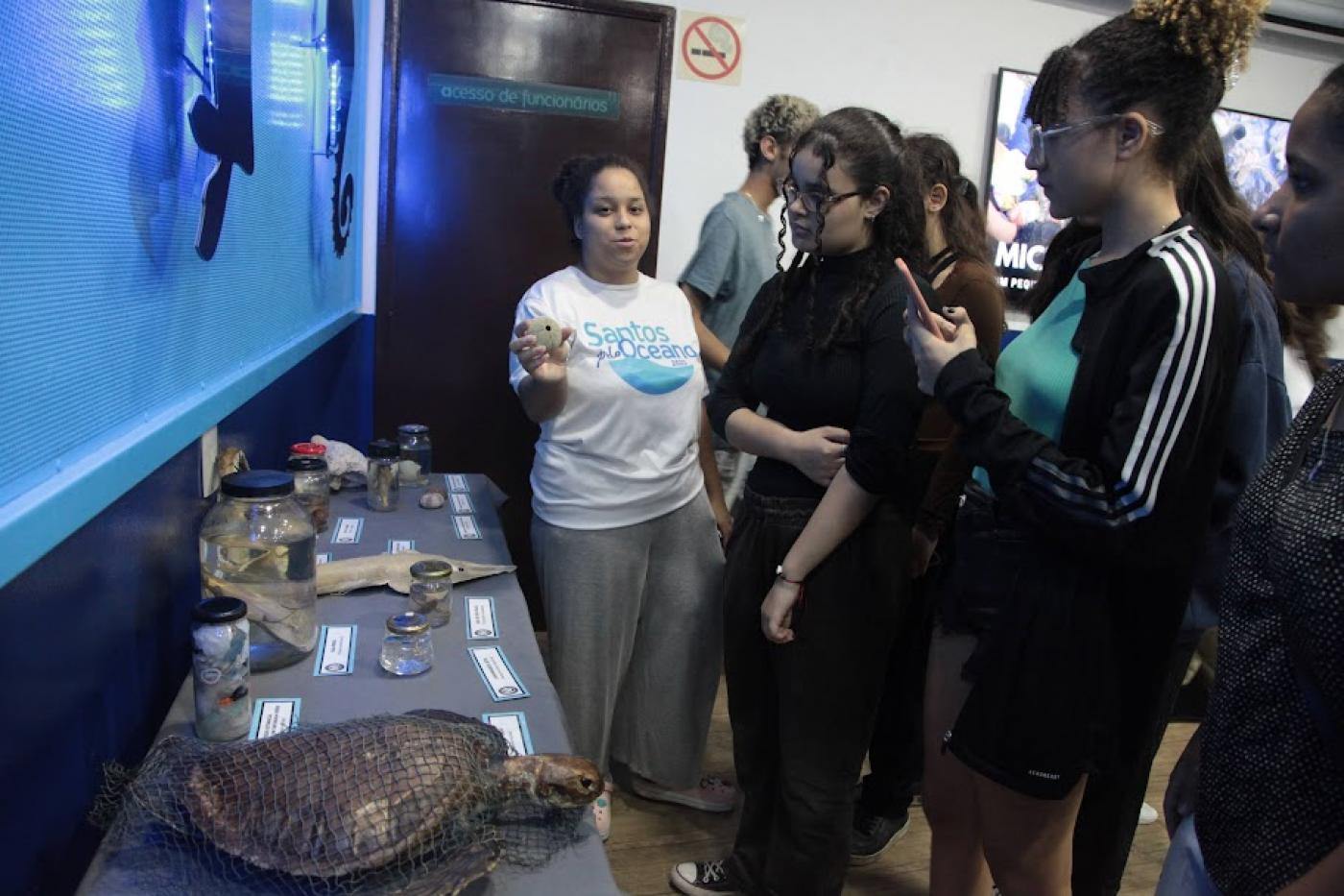 jovens estão em pé diante de mesa com animais taxidermizados. #paratodosverem