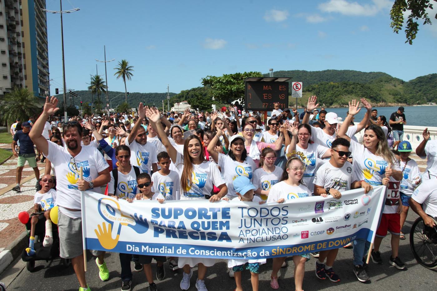 pessoas caminham com camisetas iguais e segurando uma faixa. #paratodosverem 