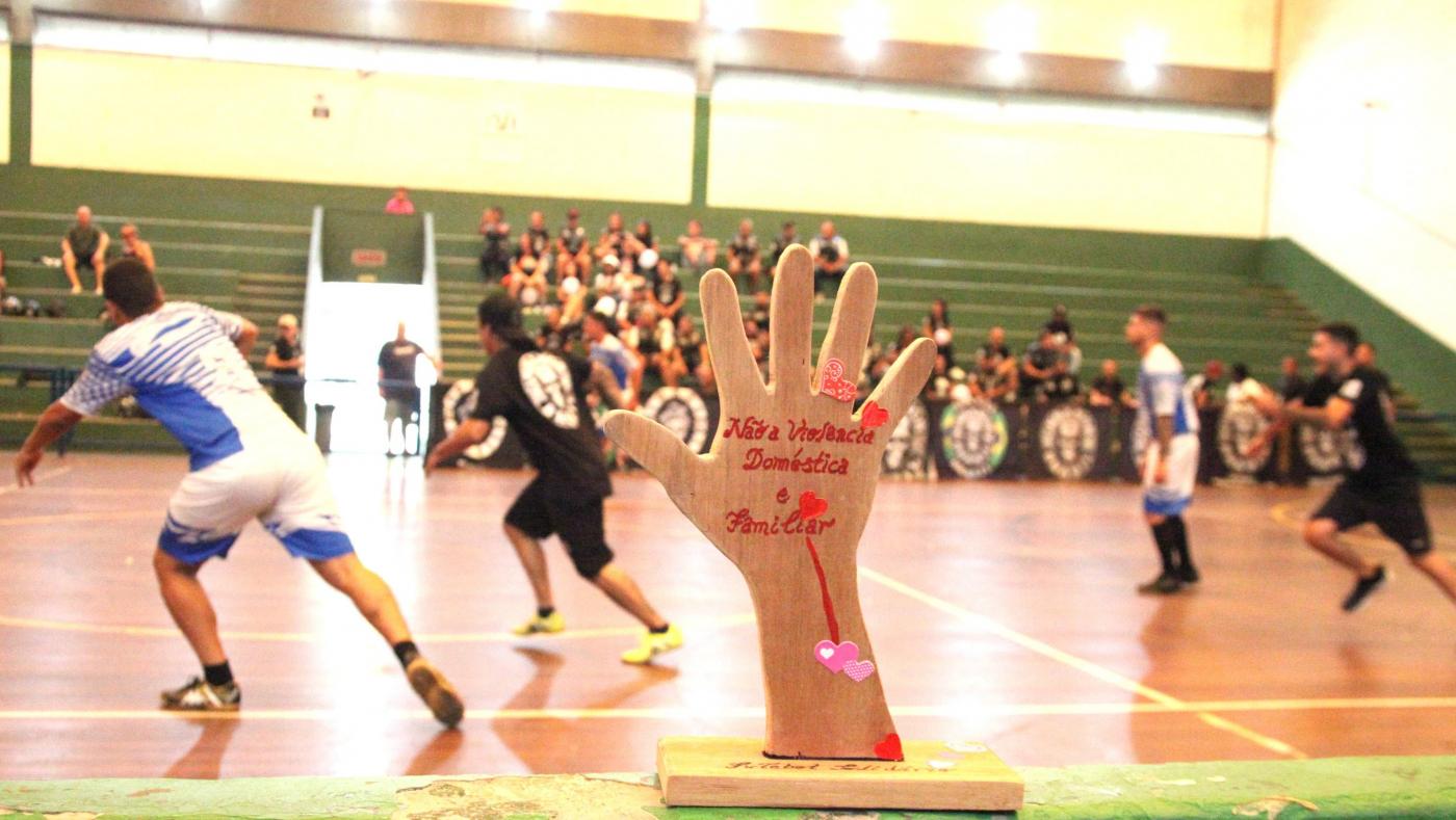 troféu com menção à mulher e jogo de futsal ao fundo. #paratodosverem