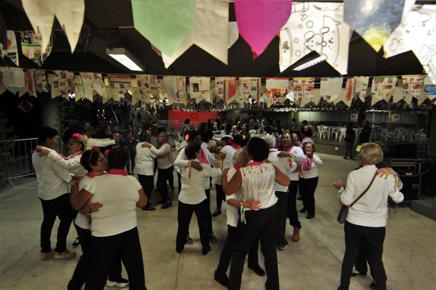 duplas de mulheres estão dançando em coreografia. Ambiente é decorado com bandeirinhas de  São João. #paratodosverem 