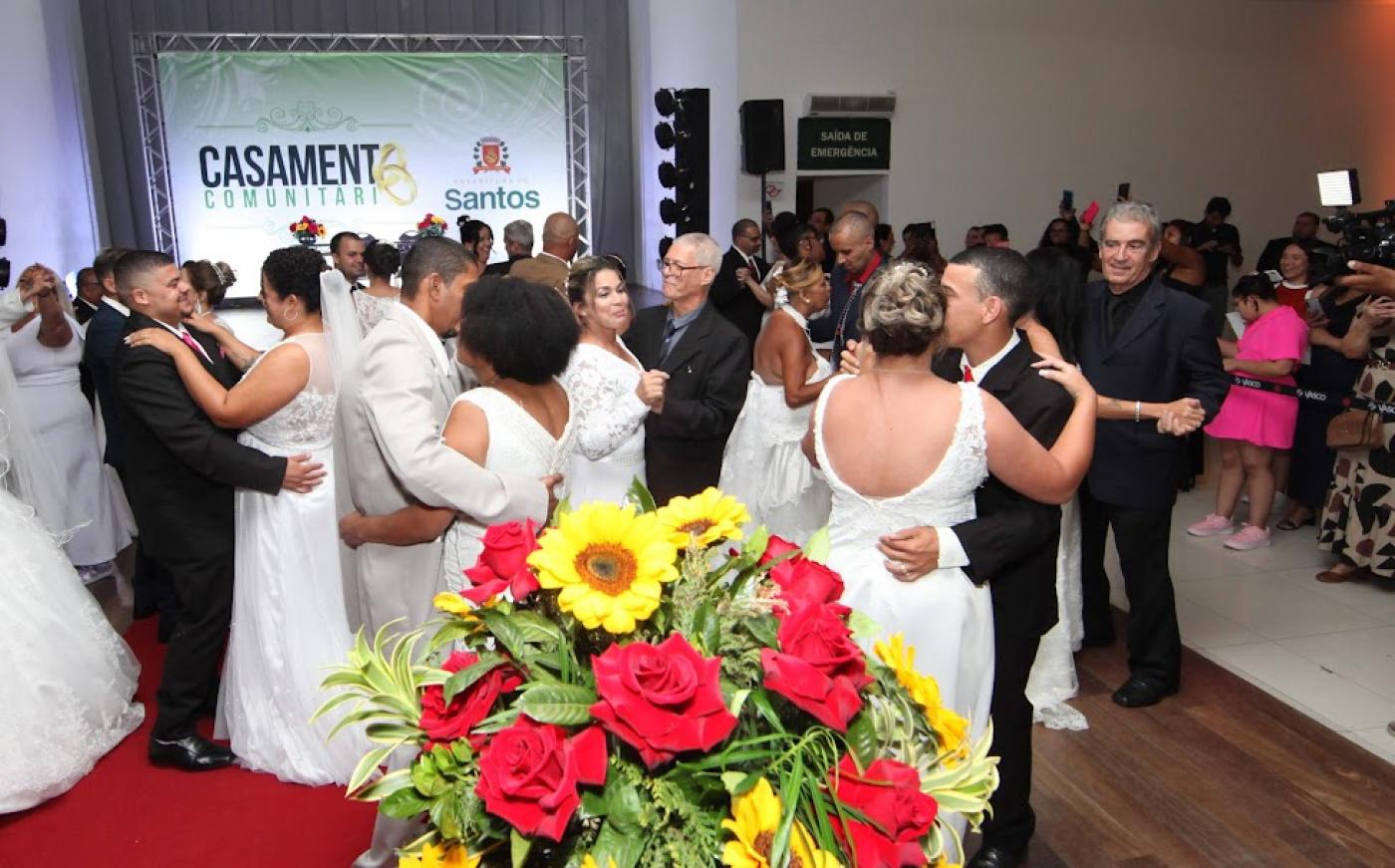 casais dançam em salão. As mulheres usam vestido de noiva e os homens, terno e gravata. Em primeiro plano há um arranjo de flores. #paratodosverem 