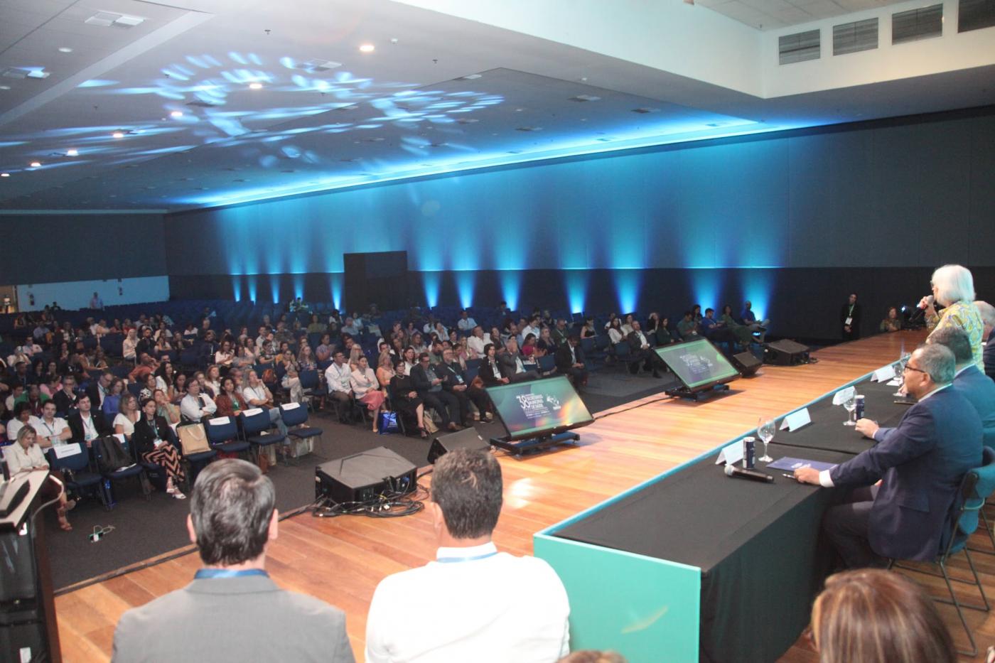 palco com mesa e pessoas no auditório lotado ao fundo. #paratodosverem 