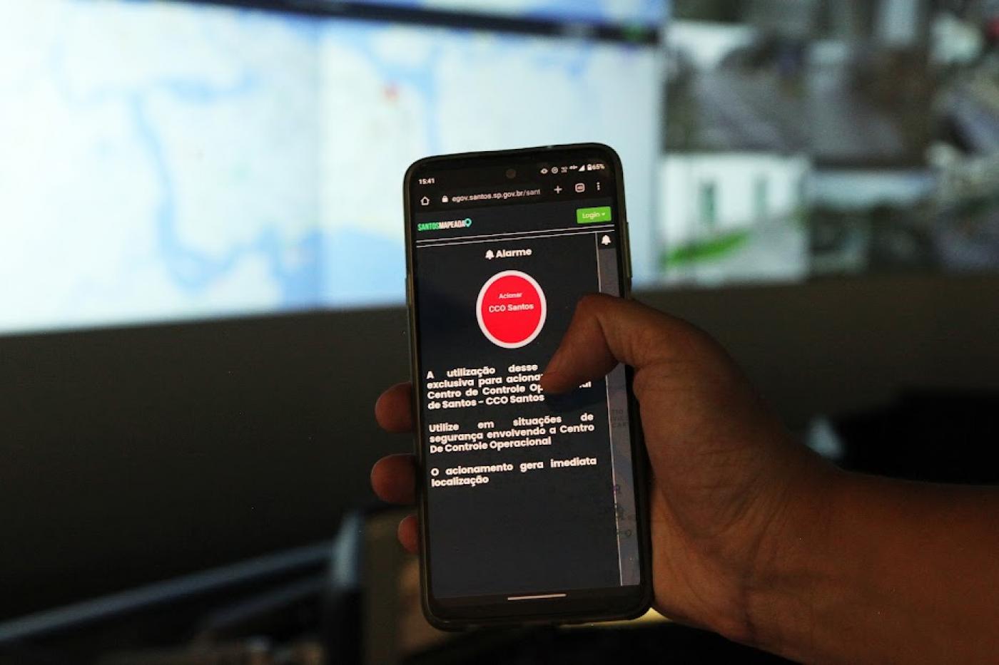 mão segura celular com botão de alerta vermelho na tela. Ao fundo, desfocado, a um mapa em um telão. #paratodosverem